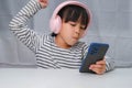 Cute elementary school girl wearing headphones holding a smartphone. Happy Asian girl studying online on smartphone or Royalty Free Stock Photo
