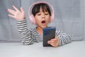 Cute elementary school girl wearing headphones holding a smartphone. Happy Asian girl studying online on smartphone or Royalty Free Stock Photo