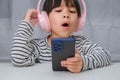 Cute elementary school girl wearing headphones holding a smartphone. Happy Asian girl studying online on smartphone or Royalty Free Stock Photo