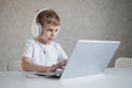 Cute european boy listens to music on headphones and types on a laptop. The child does homework on the computer. A young Royalty Free Stock Photo