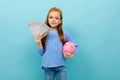 Cute european girl holding a piggy bank and money in hands on a light blue wall Royalty Free Stock Photo
