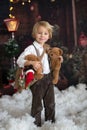 Cute fashion toddler boy, playing in the snow with teddy bear in front of a wooden cabin log Royalty Free Stock Photo