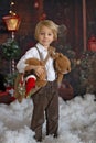 Cute fashion toddler boy, playing in the snow with teddy bear in front of a wooden cabin log