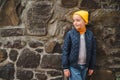 Cute fashionable boy in jacket and yellow hat, outdoors. Handsome boy at walk. Kids fashion. Happy stylish boy posing near stone Royalty Free Stock Photo