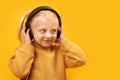 Cute flirtatious fair-haired preschool girl listens to music with headphones. Close up portrait on yellow background