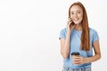 Cute friendly redhead girl inviting mate to drink coffee in cafe with her holding smartphone near ear and paper cup Royalty Free Stock Photo