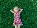 Cute funny adorable blonde Caucasian smiling girl in red pink dress lying on grass enjoying spring summer sunny day. Royalty Free Stock Photo
