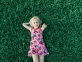 Cute funny adorable blonde Caucasian smiling girl in red pink dress lying on grass enjoying spring summer sunny day. Royalty Free Stock Photo