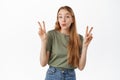 Cute and funny blond girl shows peace v-sign and pucker lips silly, standing happy in summer t-shirt and jeans against Royalty Free Stock Photo