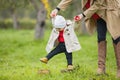 Cute funny happy baby making his first steps on a green lawn in autumn garden, mother holding his hands supporting by learning to Royalty Free Stock Photo