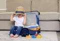 Cute funny little baby boy siiting in blue suitcase with hat on his eyes, packed for vacation full of clothes ready for traveling. Royalty Free Stock Photo