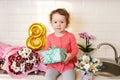 Cute funny little girl holding gift box sitting on kitchen table with bouquets of spring flowers and golden ballon 8 eight , Royalty Free Stock Photo