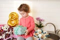 Cute funny little girl holding gift box sitting on kitchen table with bouquets of spring flowers and golden ballon 8 eight , Royalty Free Stock Photo