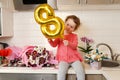 Cute funny little girl holding golden ballon 8 eight sitting on kitchen table with bouquets of spring flowers , greeting mother or Royalty Free Stock Photo