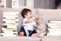 Cute funny playful little baby boy reading book and playing with teddy bear toy putting glasses on it sitting on sofa between pile Royalty Free Stock Photo