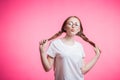 Cute funny young student girl in white t-shirt, glasses and braid shows kiss lips on a pink background with copy space