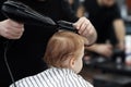 Cute ginger baby boy in a barber shop having haircut by hairdresser. Hands of stylist with tools hair dryer and hairbrush.