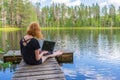 Cute ginger girl sitting on wooden planked footway and working with laptop in summer day against beautiful landscape of northern Royalty Free Stock Photo