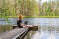 Cute ginger girl sitting on wooden planked footway and working with laptop in summer day against beautiful landscape of northern Royalty Free Stock Photo