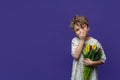 Cute girl in beige casual dress holds armful of tulips and laughs by closing her mouth with her hand on purple background. Copy