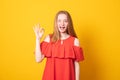 Cute girl blinks eye, makes okay gesture, demonstrates her agreement, feels happy, wears red dress