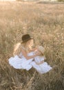 Cute girl with blond long hair with a younger sister in a summer field at sunset with a white dress with a straw hat