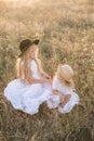 Cute girl with blond long hair with a younger sister in a summer field at sunset with a white dress with a straw hat