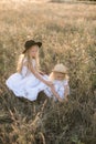 Cute girl with blond long hair with a younger sister in a summer field at sunset with a white dress with a straw hat