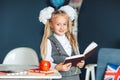 Cute girl blondy in school uniform with white bows standing near the desk and studying in the classroom. Education and school