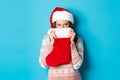 Cute girl cover face with christmas stocking, staring right with cunning gaze, standing in Santa hat and celebrating