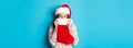 Cute girl cover face with christmas stocking, staring right with cunning gaze, standing in Santa hat and celebrating