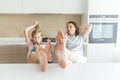 Cute girl and her mother are smiling while eating ice cream in the kitchen with legs on a table