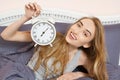 Cute girl holds a clock and lies in bed bedroom. Emotional beautiful woman lying in bed on her day off Royalty Free Stock Photo