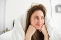 Cute girl with messy hair, lying in bed covered in white sheets duvet, smiling and laughing coquettish, spending time in Royalty Free Stock Photo
