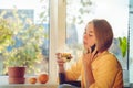 Cute woman with a mole on her cheek talking on the phone sitting near the window with a mug of tea