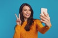 Cute girl in orange sweater showing peace gesture while taking selfie with smartphone Royalty Free Stock Photo