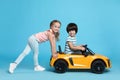 Cute girl pushing children`s electric toy car with little boy on light blue background