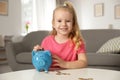 Cute girl putting coin into piggy bank at table in living room. Royalty Free Stock Photo