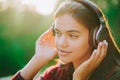 Cute girl`s face with headphones. Young woman enjoys music while sitting in green park. Concept of youth, freedom