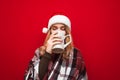 Cute girl in Santa hat and plaid drinks hot drink from big cup with closed eyes and enjoys on red background. Lady drinks tea and Royalty Free Stock Photo