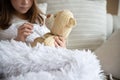 A cute girl is sick under a blanket on the sofa and measures the temperature of the teddy bear.