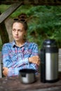 Cute girl sitting on a wooden table with thermos with hot drink and gets her hands warm. Female person feels cold and trying to