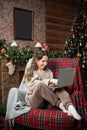 A cute girl in a sweater is sitting in a chair near the Christmas tree and looking at a laptop Royalty Free Stock Photo