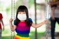 Cute girl wear white medical face mask to prevent the spread of coronavirus COVID-19. Little child sat on playground swing. Royalty Free Stock Photo