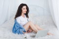 A cute girl in a white T-shirt and denim shirt, sits on a bed and uses a laptop. Royalty Free Stock Photo