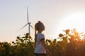 Cute girl in white t-shirt smelling sunflower in sunset field wind turbines farm on background. Child with long braid Royalty Free Stock Photo