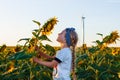 Cute girl in white t-shirt smelling sunflower in sunset field wind turbines farm on background. Child with long braid Royalty Free Stock Photo