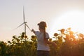Cute girl in white t-shirt smelling sunflower in sunset field wind turbines farm on background. Child with long braid