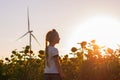 Cute girl in white t-shirt smelling sunflower in sunset field wind turbines farm on background. Child with long braid Royalty Free Stock Photo