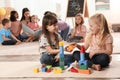 Cute girls playing with building blocks on floor while kindergarten teacher reading book to other children Royalty Free Stock Photo